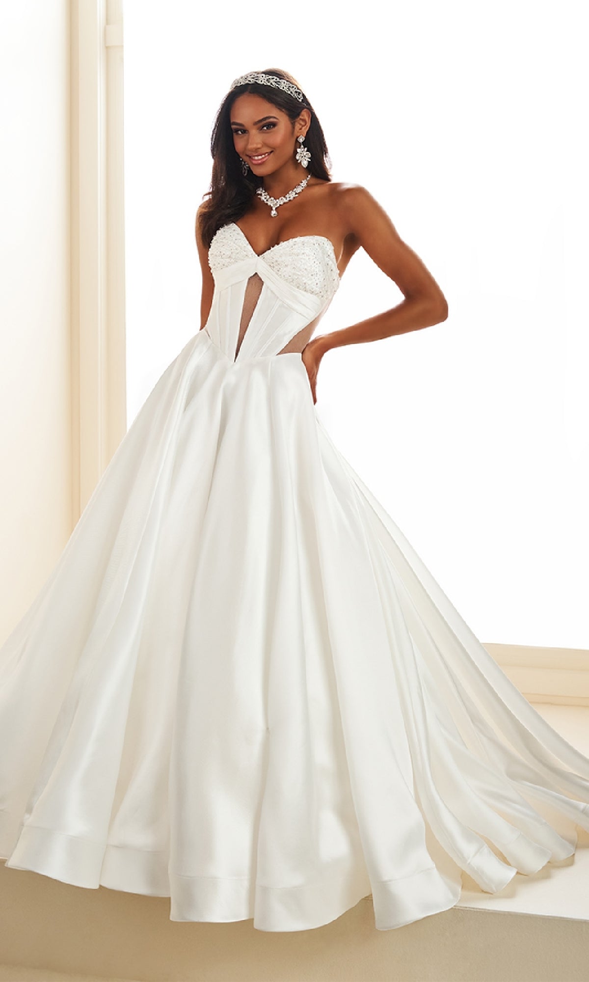 A woman stands indoors wearing the Ellie Wilde EW36025, a strapless white satin prom ball gown with a corset bodice and full skirt, styled with statement earrings and a tiara.